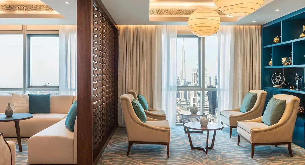 A modern high-rise hotel suite with a spacious seating area, cream armchairs with teal pillows, a blue accent wall with decorative objects, a coffee table, large windows with city views, and curtains.