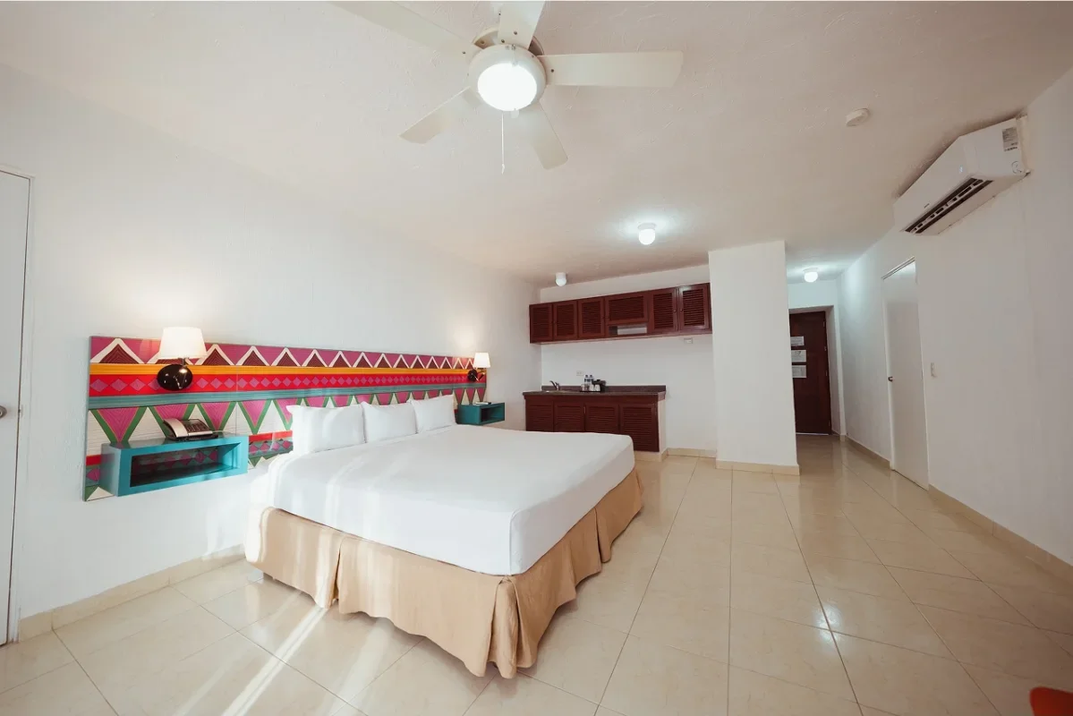 Hotel room with a large bed, white bedding, colorful headboard, wall-mounted lamps, ceiling fan, air conditioning unit, and a kitchenette with wooden cabinets.