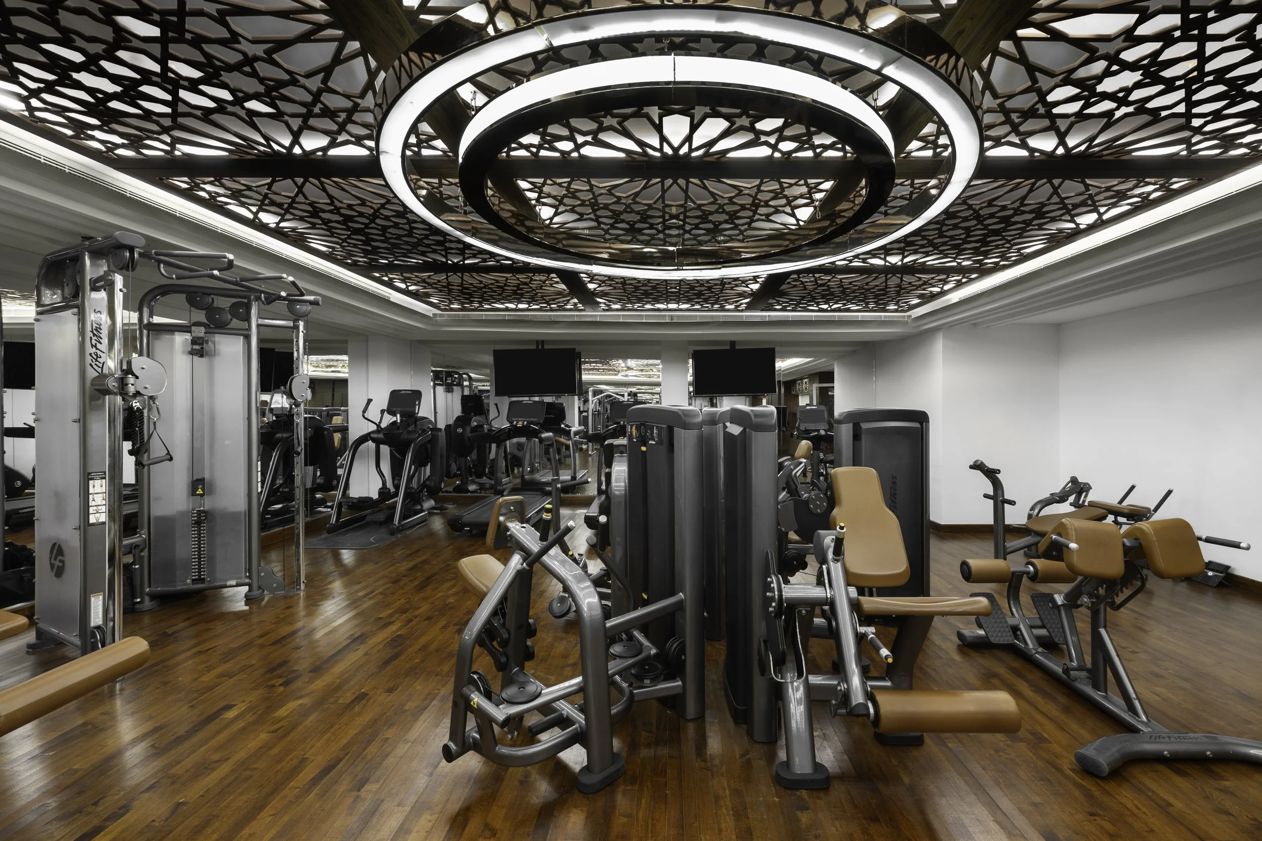 Interior of a modern gym with various workout equipment and a decorative ceiling featuring intricate geometric patterns and circular light fixtures.