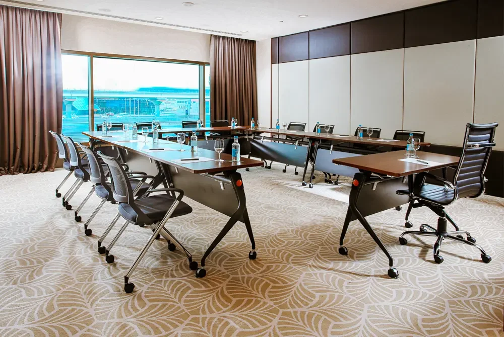 A modern meeting room with a U-shaped conference table, black rolling chairs, and large windows with brown curtains.