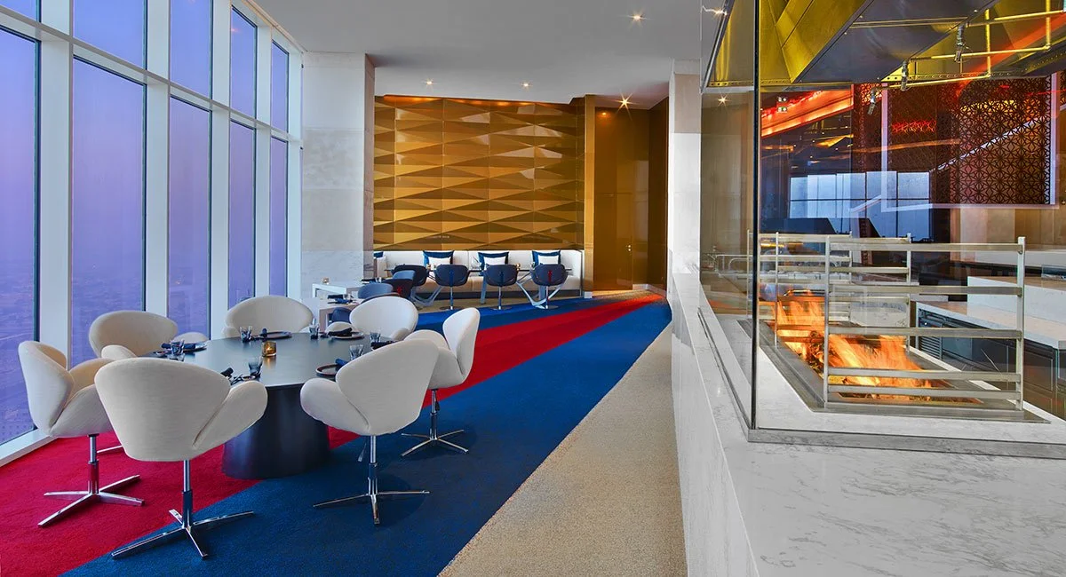 Modern dining area with white chairs around a black round table, large windows with blue tint, colorful carpet, and a fireplace with glass enclosure.