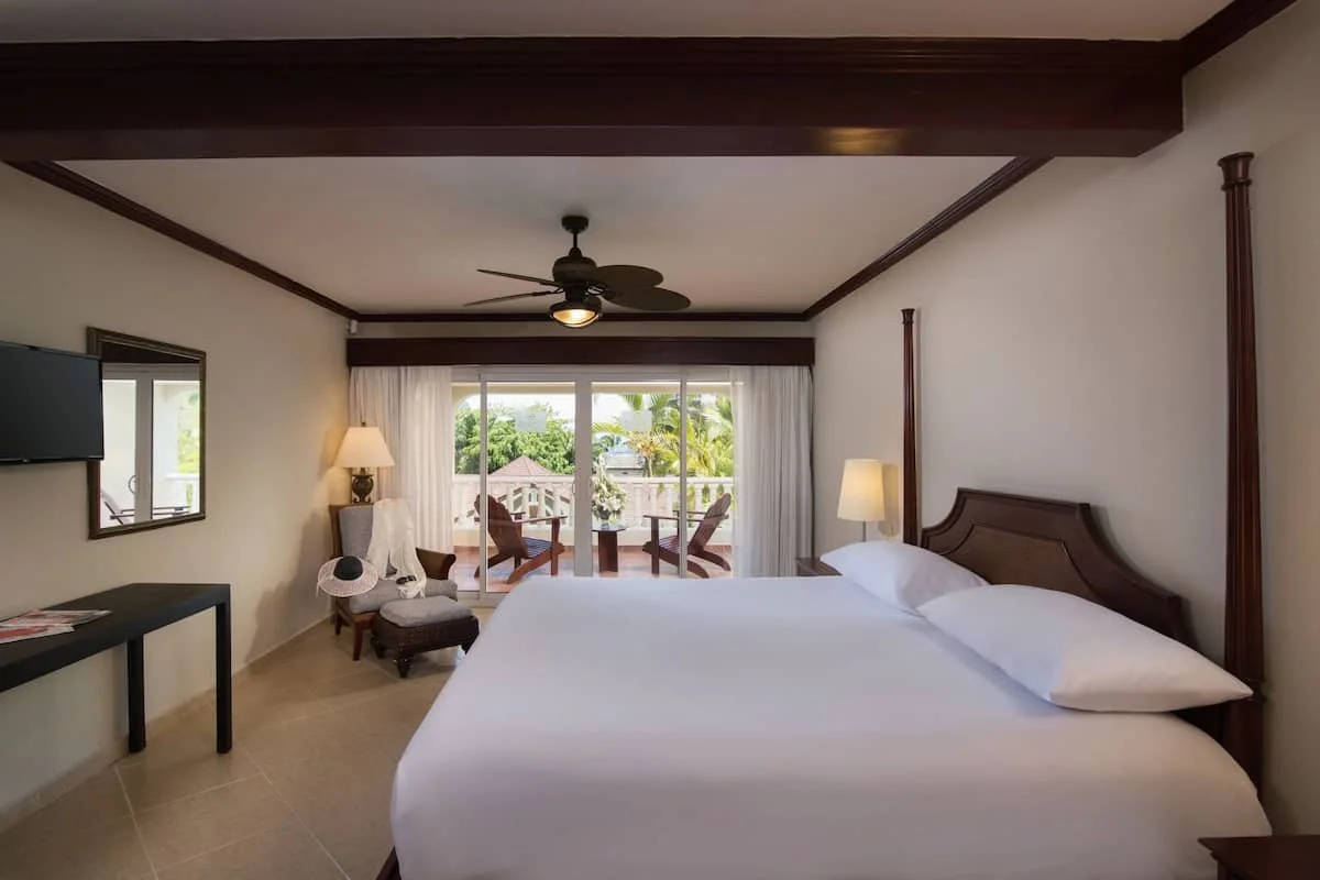 Hotel room with a large bed, a ceiling fan, a balcony with outdoor chairs, a television, and side tables with lamps.