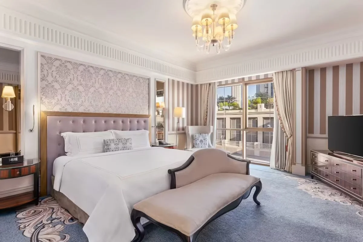Elegant hotel room with a large bed, tufted headboard, patterned wallpaper, armchair, TV, and large window with curtains.