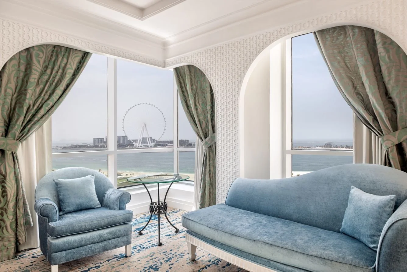 Living room with large window view of a city skyline, Ferris wheel, and water, decorated with blue velvet armchair, matching sofa, and patterned curtains.