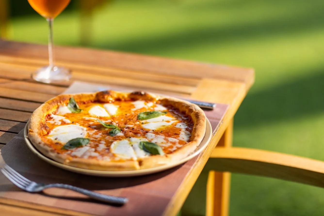 Pizza with cheese, tomato sauce, and basil leaves on a wooden table outdoors with a glass of beverage in the background.
