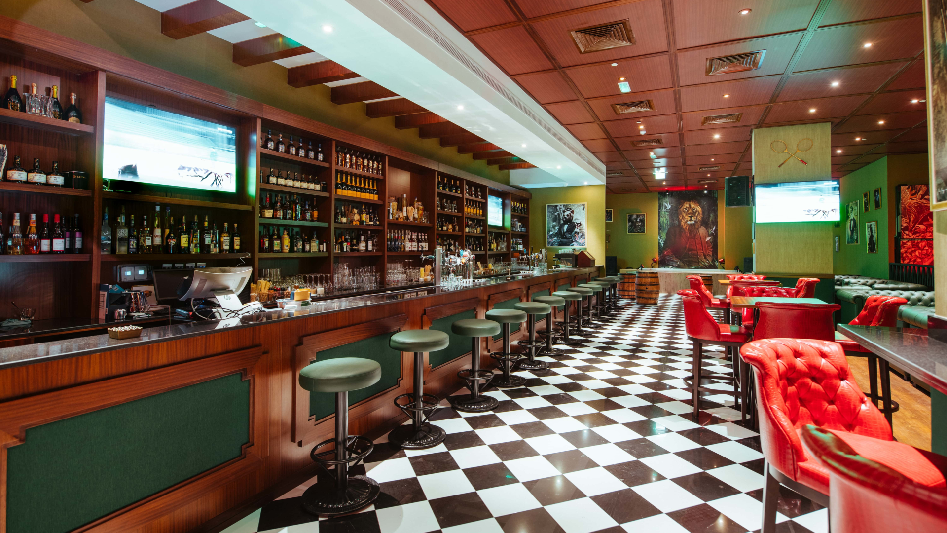 Interior of a bar and lounge with a checkered black and white floor, wooden bar counter, green and red upholstered chairs, and wall art including a lion portrait and tennis rackets.