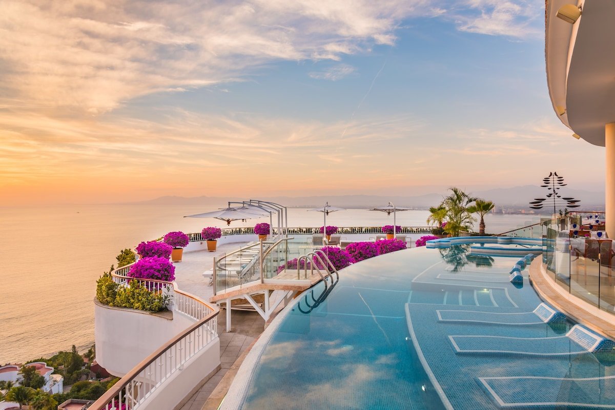 Luxury rooftop infinity pool overlooking a sunset over the ocean, with pink flowering plants, white umbrellas, and palm trees.