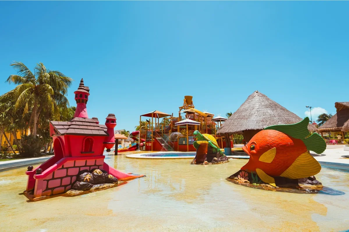 Colorful children's water play area with a pink castle, a large fish sculpture, and a turtle sculpture, set under blue skies with palm trees and thatched-roof structures.
