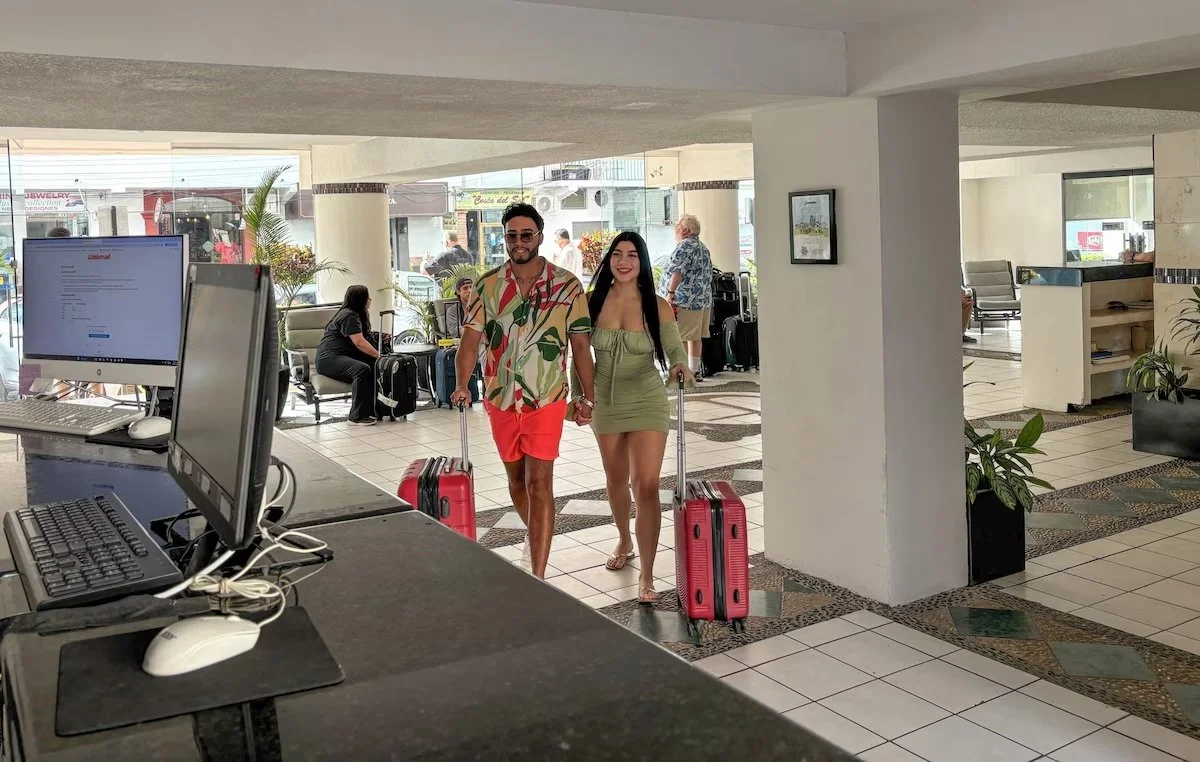A smiling man and woman holding hands and pulling pink suitcases walk through a hotel lobby entrance, with other travelers and seating visible in the background.