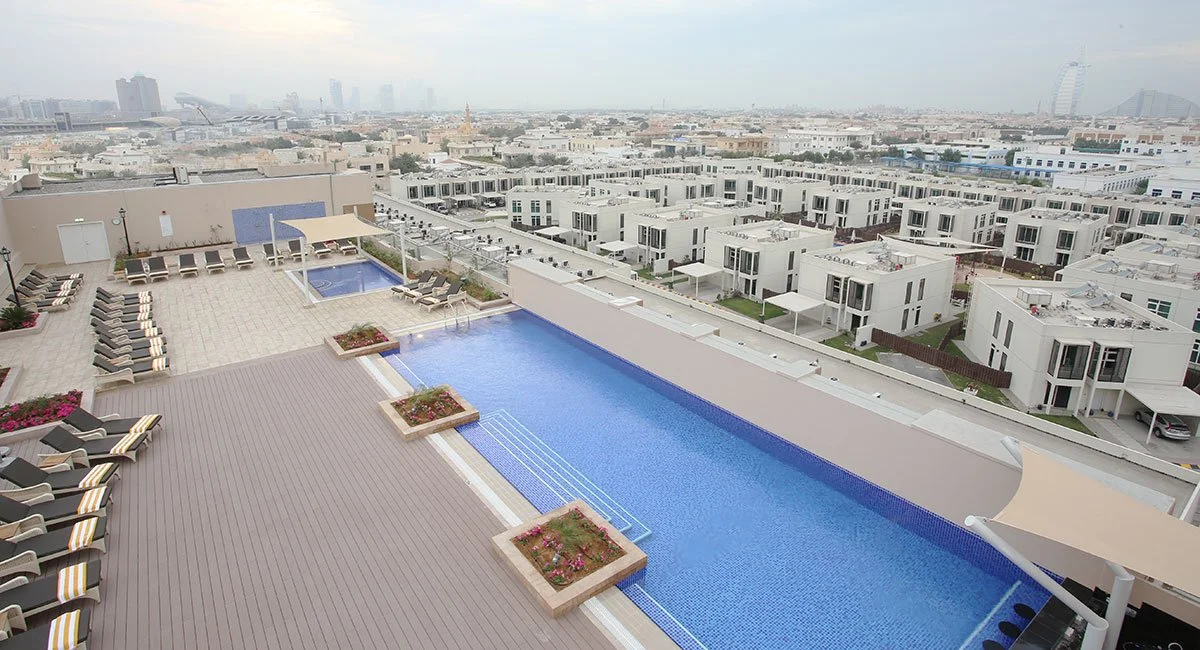 Rooftop swimming pool with lounge chairs and planters, overlooking a cityscape with modern residential buildings and a distant skyline.