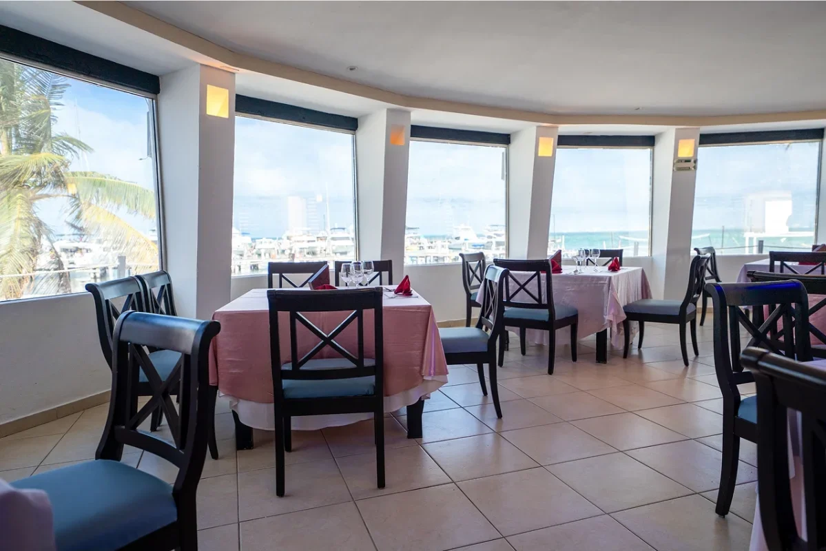 Interior of a restaurant with tables covered in pink tablecloths, set with glasses and red napkins, overlooking a marina through large windows.