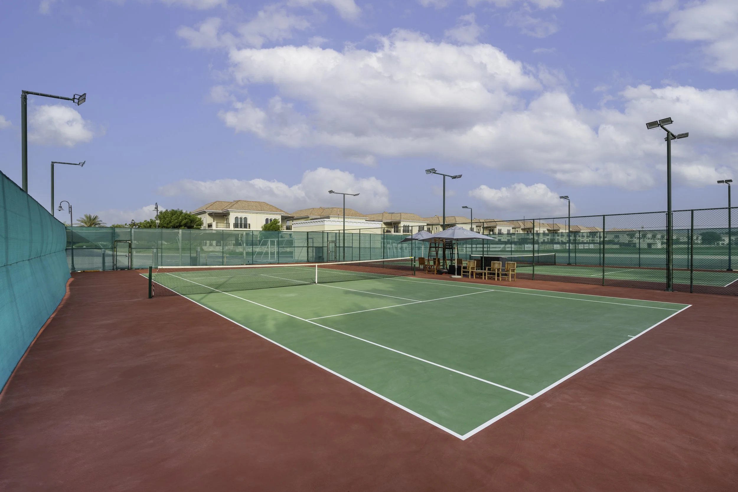 Empty outdoor tennis court with green and red surface, surrounded by green fencing, under a partly cloudy blue sky with residential houses in the background.