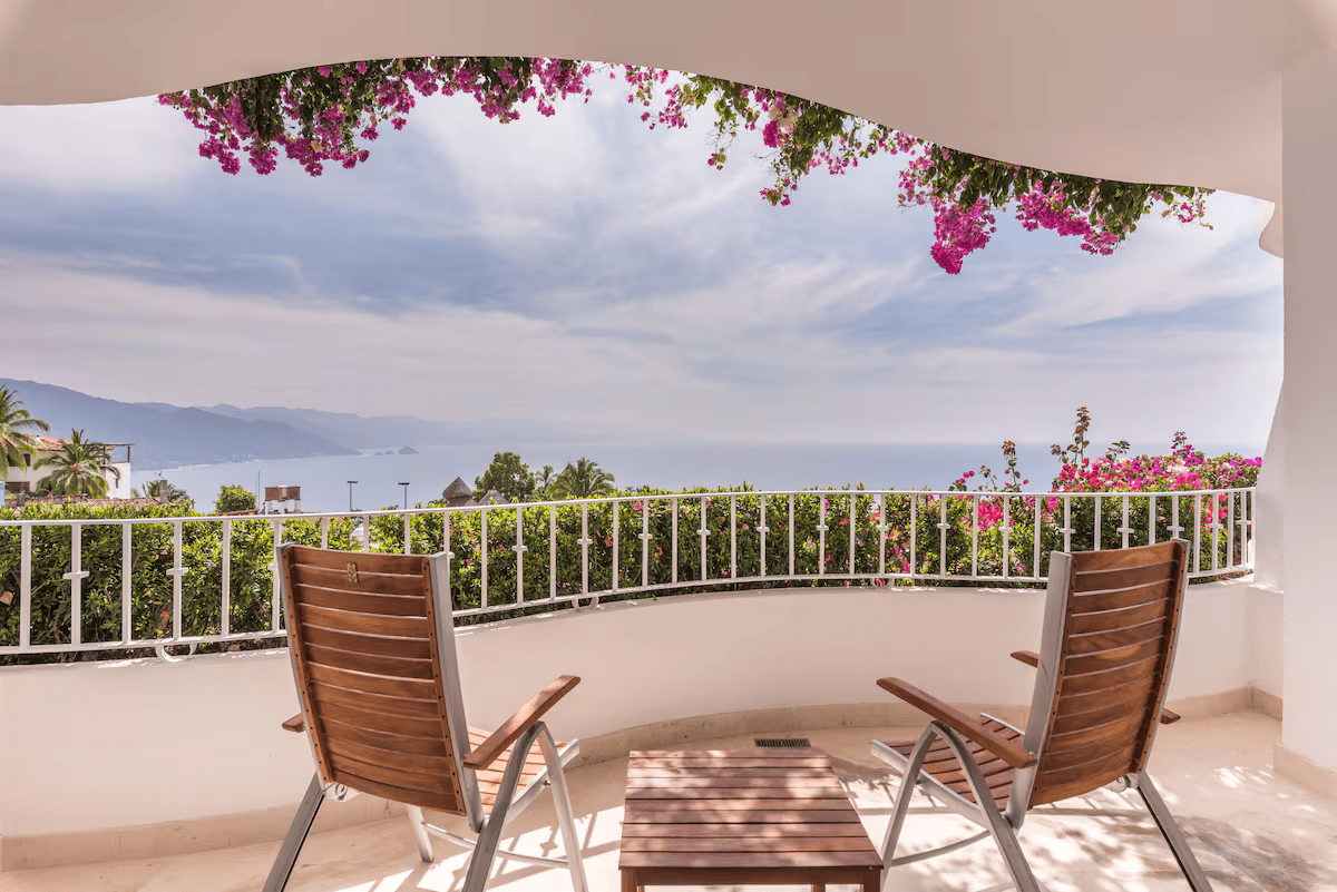 Balcony with two wooden chairs and a small table overlooking a lush landscape, flowers hanging overhead, and mountains in the distance under a cloudy sky.