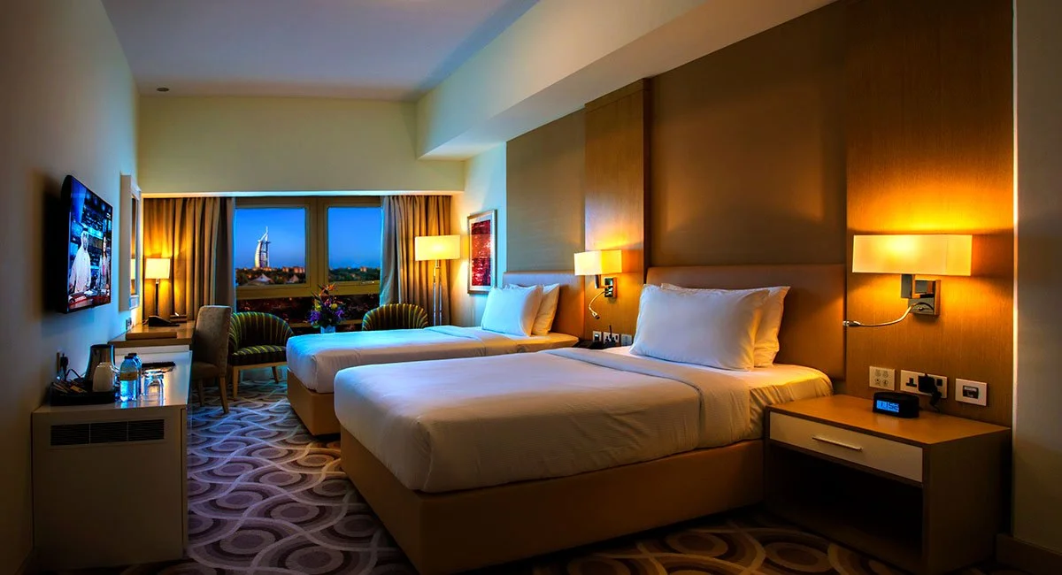 A hotel room with two twin beds, a window with curtains, a sitting area with two chairs and a small table, and a mounted flat-screen TV.