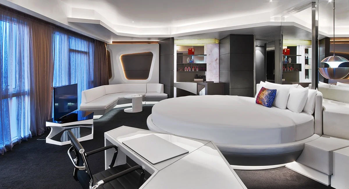 Modern hotel room with a large circular bed, white sofa, flat-screen TV, desk with chair, and large windows with blue curtains.