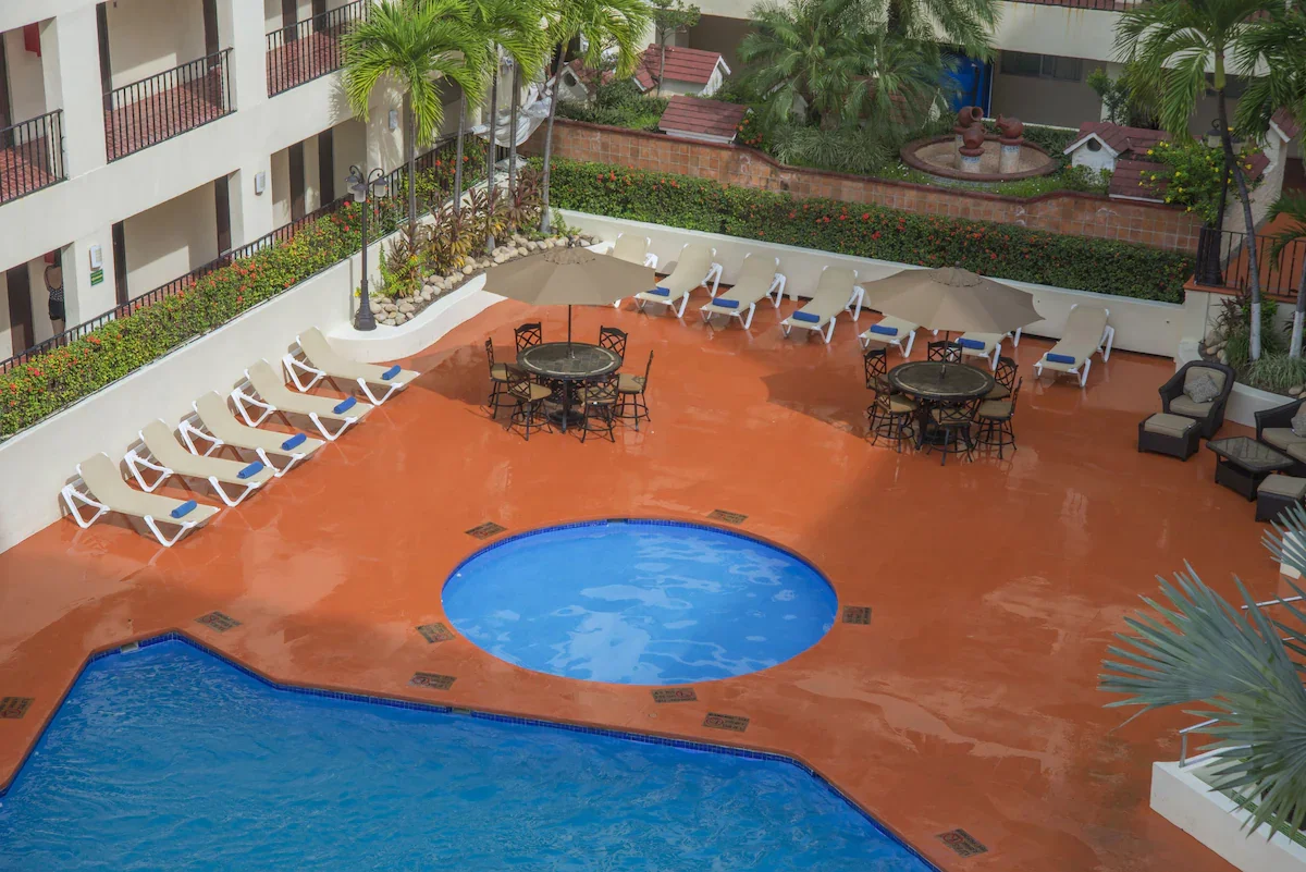 Outdoor swimming pool area with multiple lounge chairs, tables, umbrellas, and palm trees.
