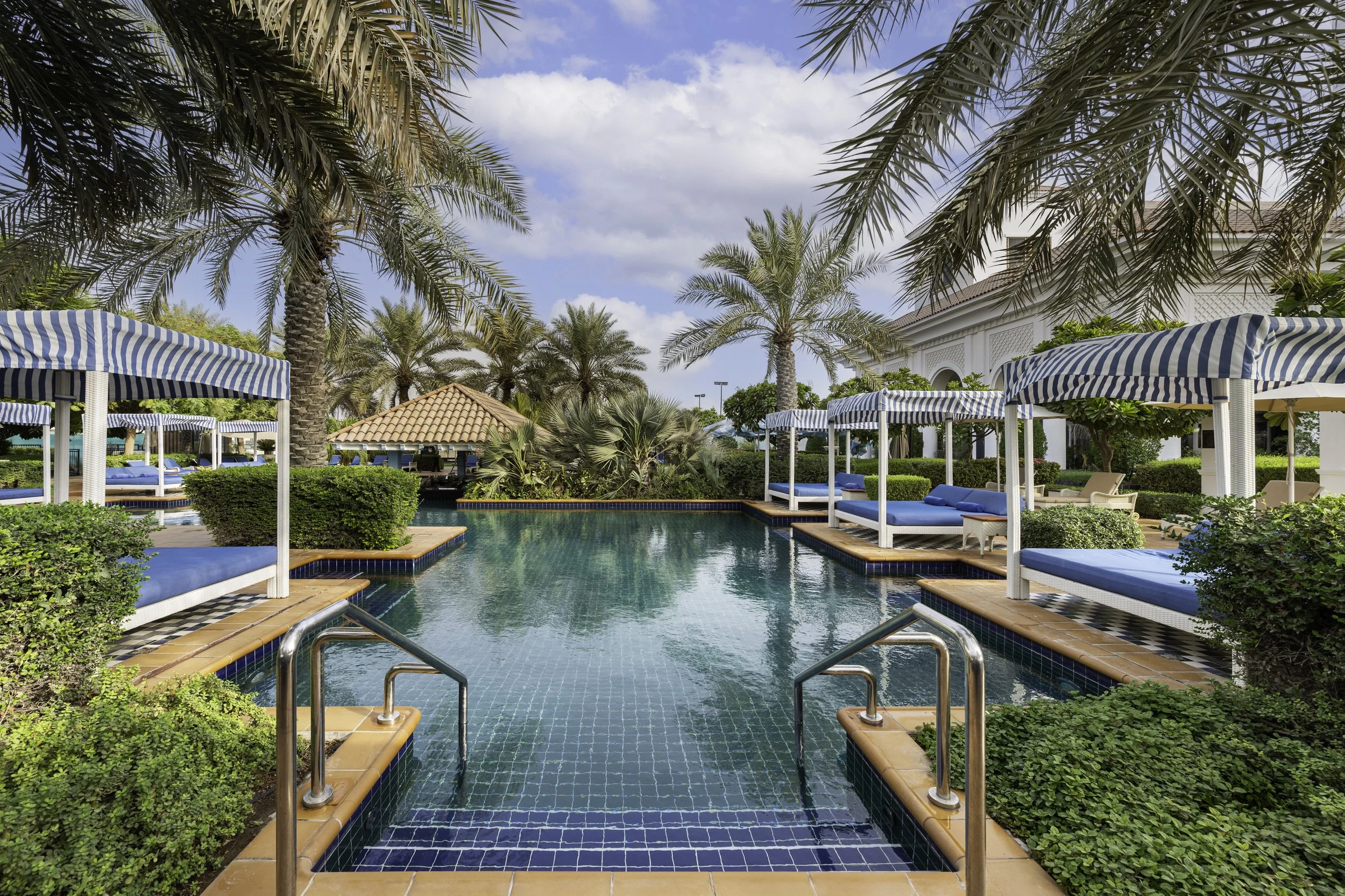 A luxurious pool area surrounded by palm trees, with lounge chairs covered by blue and white striped umbrellas, and a white building with an ornate facade in the background, under a partly cloudy sky.