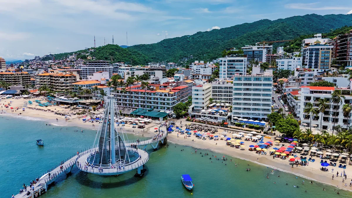 Aerial view of a beach with colorful umbrellas, a pier with a viewing platform, boats in the water, and a city with tall buildings against green hills in the background.