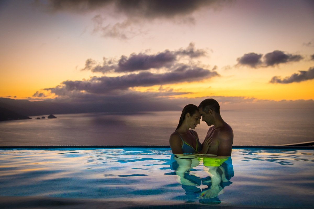 A couple in swimwear standing in a pool at sunset, with a view of the ocean and a cloudy sky in the background.