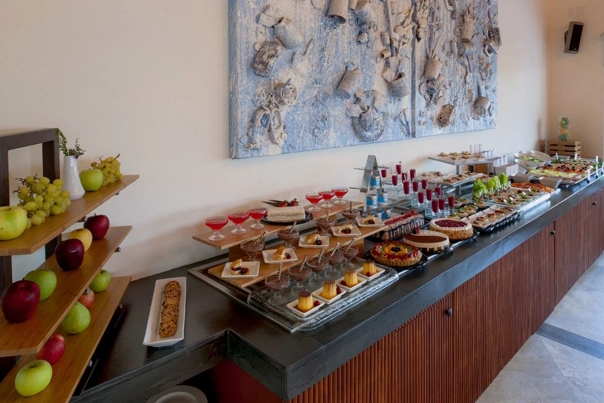 A dessert buffet with various cakes, pudding, and drinks, with some apples and grapes on a side shelf.