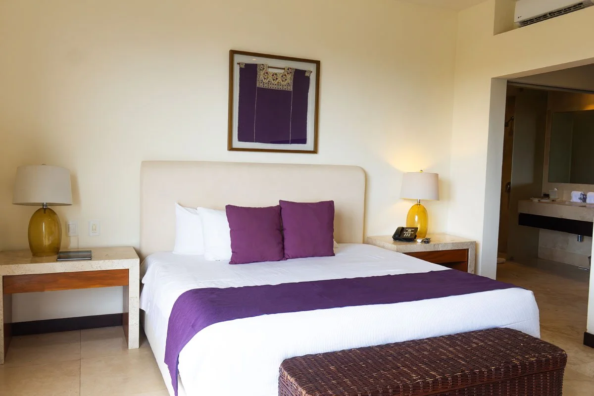 A neatly made hotel room bed with white linens, purple pillows, and a matching purple throw. There are two bedside tables with yellow lamps and a framed picture hanging above the bed. A wicker bench is at the foot of the bed, and there is a doorway t