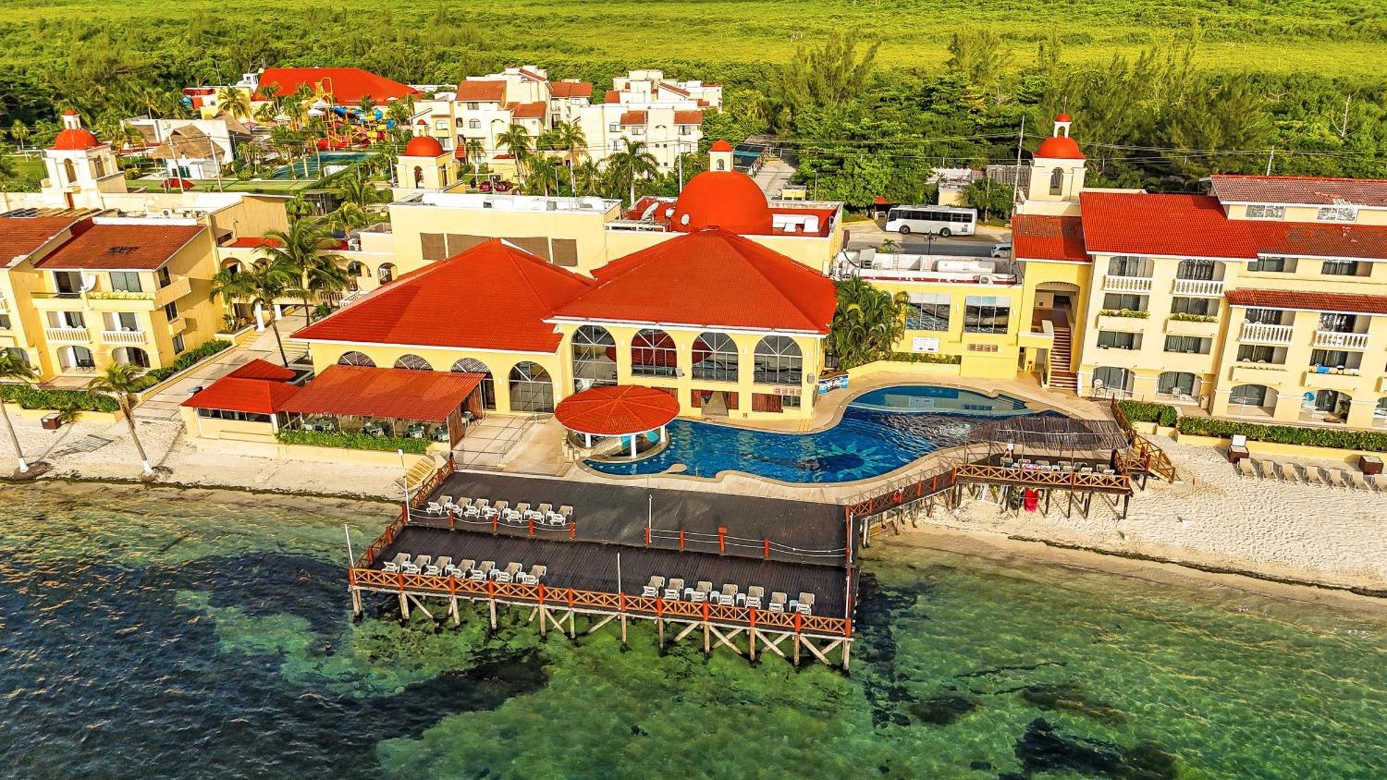 Aerial view of a resort with multiple white buildings with red roofs, a swimming pool, and a beachfront with lounge chairs, located by the ocean, surrounded by green trees and hills.