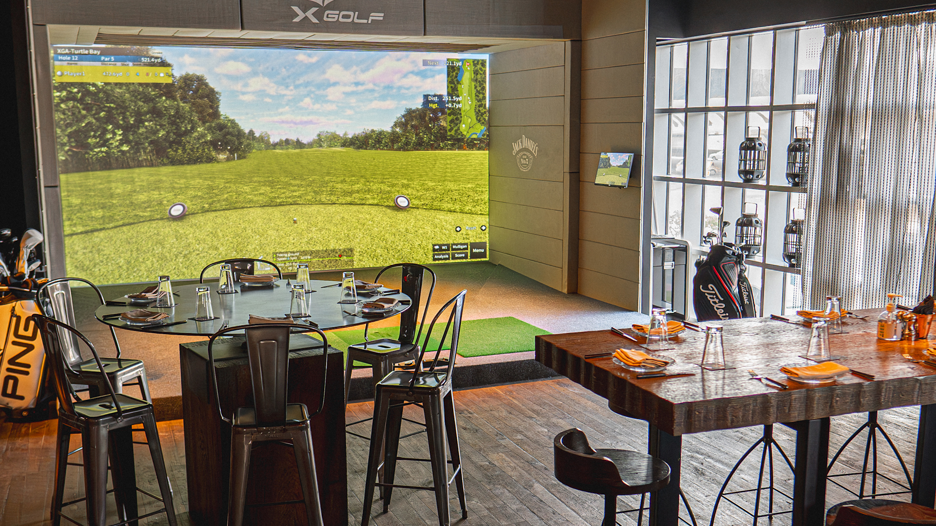 Indoor golf simulation with tables set for dining, large screen showing a virtual golf course, and golf equipment in the room.
