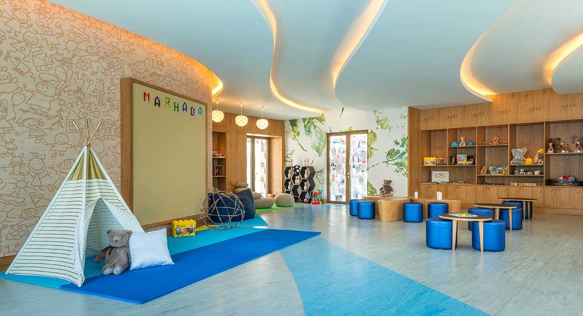 A spacious, well-lit children's playroom with a cozy teepee, toys, and soft seating areas, featuring wooden shelves and colorful decorations.
