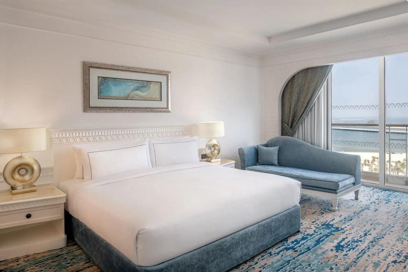 A hotel room with a large bed, two bedside lamps, a blue chaise lounge with a cushion, and a floor-to-ceiling window showing an ocean view.