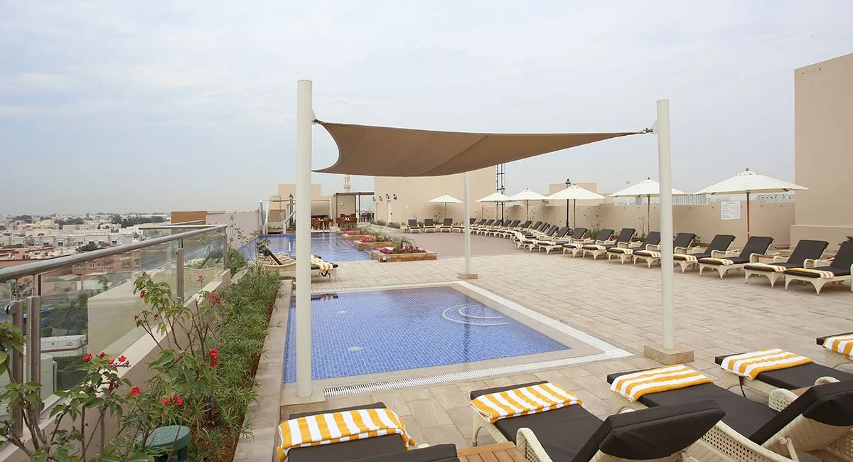 Rooftop pool area with lounge chairs, umbrellas, and a shaded section, overlooking city buildings.