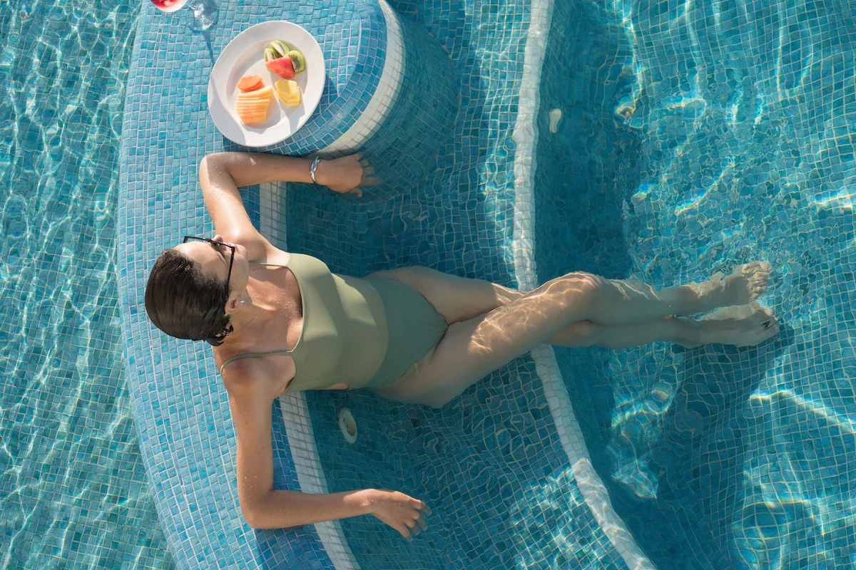 A woman in a green swimwear relaxing in a swimming pool with her feet submerged, holding a tray of sliced fruits, including watermelon, kiwi, and pineapple.