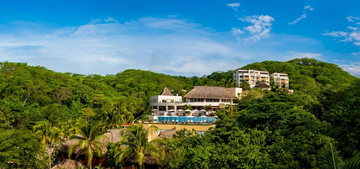 Luxury resort on a hillside surrounded by lush green trees under a blue sky with scattered clouds.
