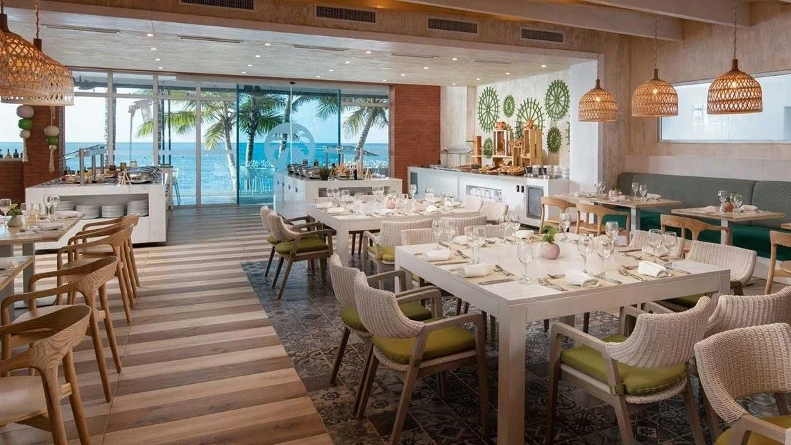 Restaurant with ocean view, decorated with hanging wicker lights, green wall art, and a variety of tables and chairs set for dining.