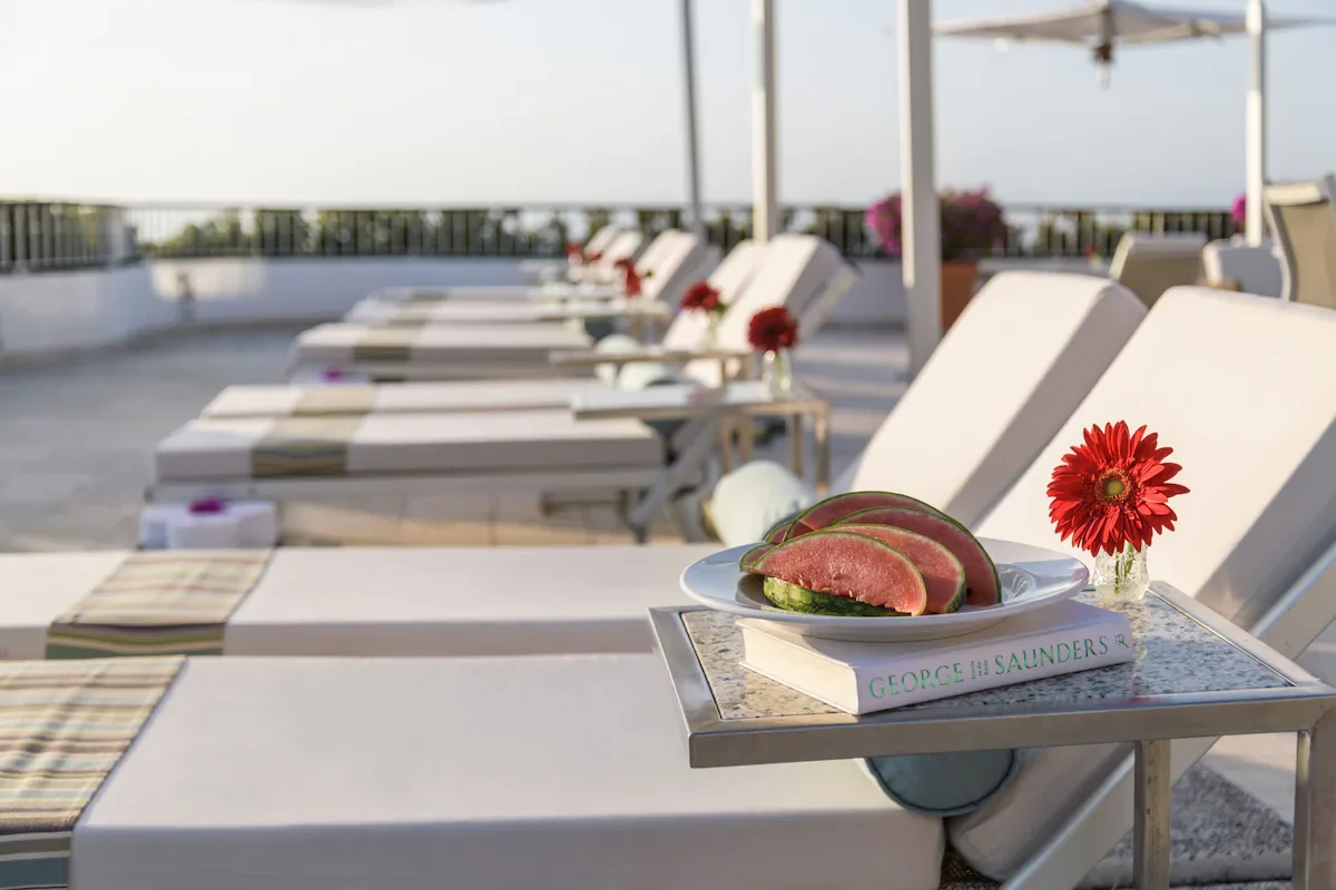 Outdoor poolside lounge chairs with side tables, each holding a book and flower vase, in a sunny setting.
