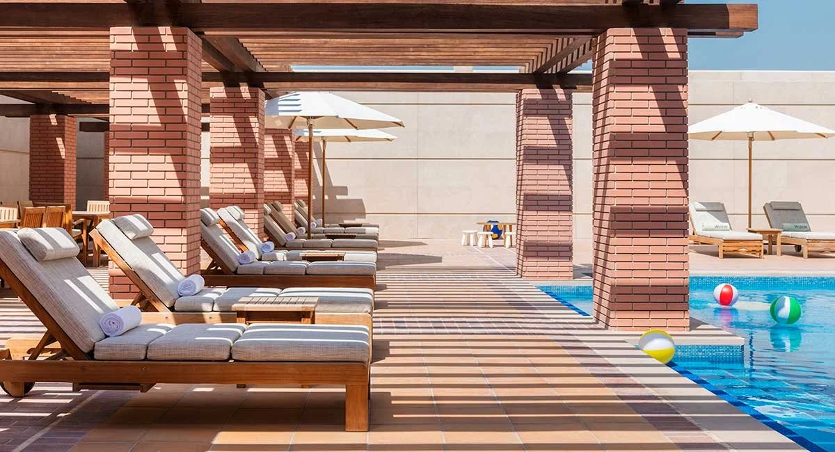 Empty poolside lounge area with wooden chairs, umbrellas, and a swimming pool with toys.