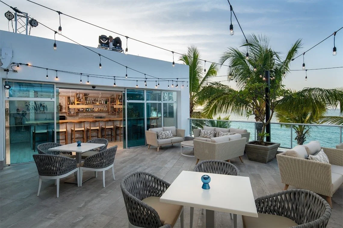 Outdoor rooftop bar and lounge area with white and grey seating and string lights, overlooking the ocean with palm trees.
