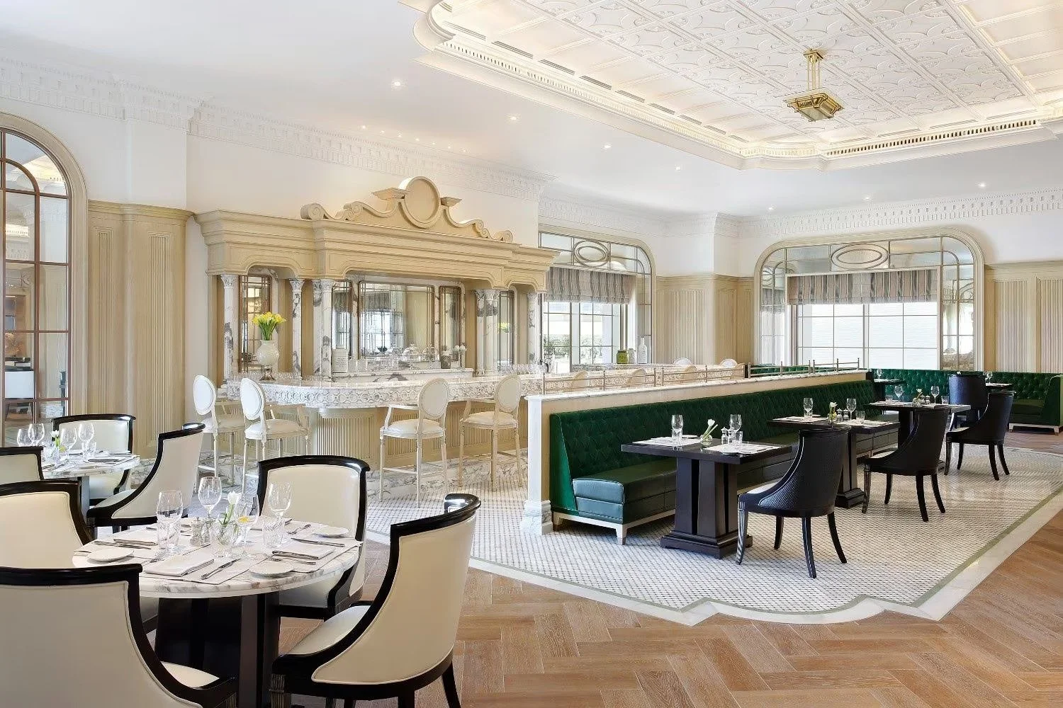 Elegant restaurant interior with white walls, large windows, green velvet booths, and round tables set with glassware and cutlery.