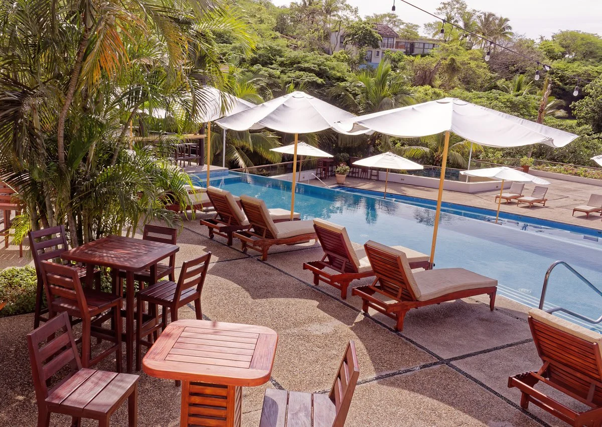 Swimming pool surrounded by lounge chairs with umbrellas, outdoor tables and chairs, lush greenery, and trees in a residential area.