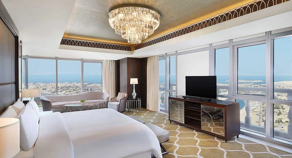 Luxury hotel room with large windows showing city and ocean views, king-sized bed, chandelier, flat-screen TV, sofa, and modern decor.