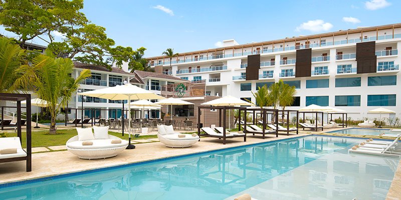 Luxury hotel pool area with lounge chairs, umbrellas, and palm trees outside a modern hotel building under a sunny sky.