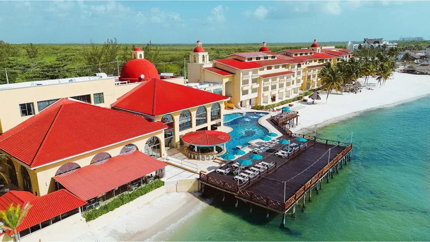 Aerial view of a luxury beachfront resort with yellow buildings featuring red roofs, a swimming pool, and a wooden pier extending into the ocean, along a sandy beach with umbrellas and palm trees.