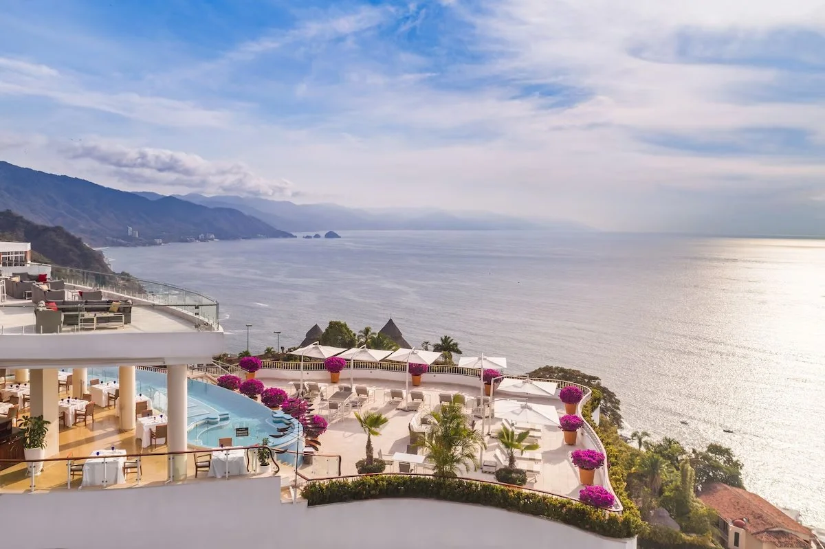 View of a luxury hotel terrace overlooking the ocean with potted pink flowers, umbrellas, and seating areas, mountains in the background under a partly cloudy sky.