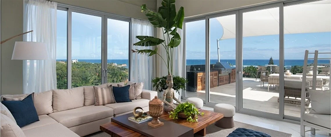 Living room with large glass sliding doors opening to balcony with outdoor dining and ocean view; beige sectional sofa, blue pillows, wooden coffee table with decorative items, indoor plants.
