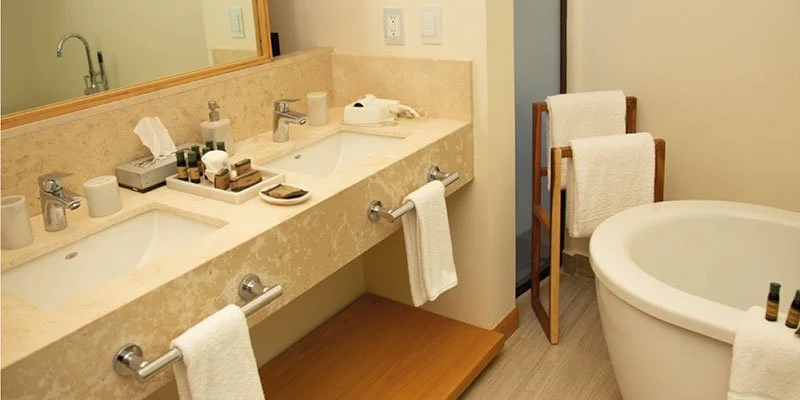 Bathroom with beige marble countertop, double sink, towels, toiletries, and a white bathtub with towels on the side.
