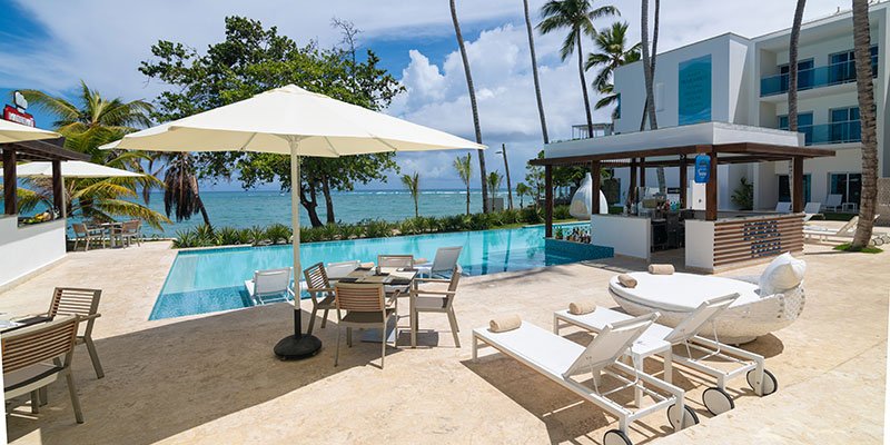 Poolside area at a tropical resort with lounge chairs, a large umbrella, and a bar, overlooking the ocean with palm trees.