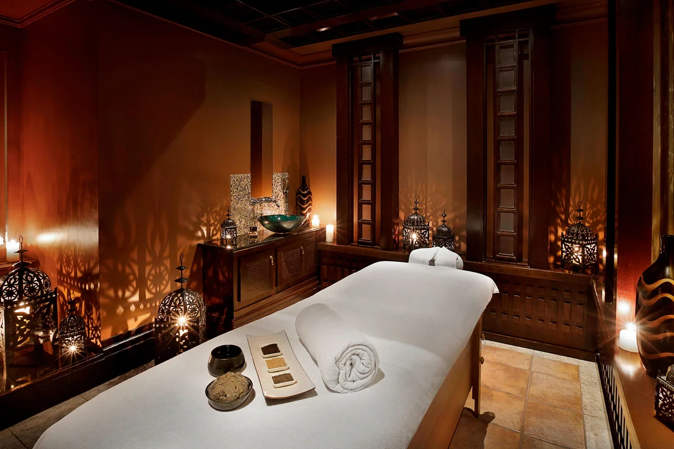 A luxurious massage room with a massage table in the center, decorated with candles, lanterns, and decorative vases, creating a relaxing atmosphere with warm lighting and Thai-inspired design elements.