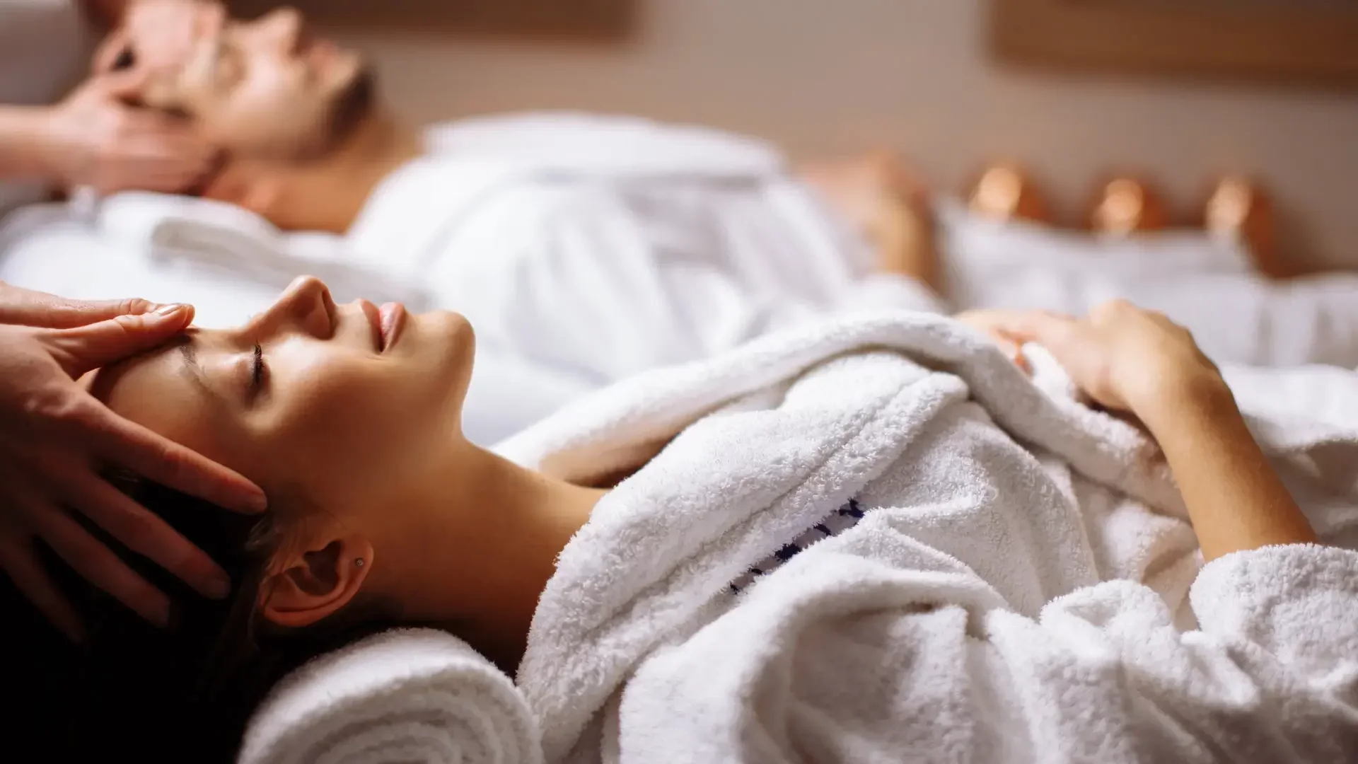 A woman is receiving a facial massage while lying in a spa or massage room. She is wrapped in a towel and appears relaxed with closed eyes.