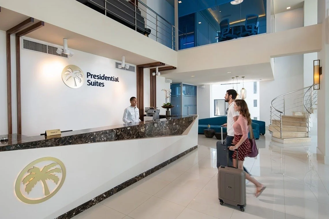 A modern hotel lobby with a reception desk labeled 'Presidential Suites', two guests with rolling suitcases checking in, and a spiral staircase. The decor is white with wooden accents and blue seating.