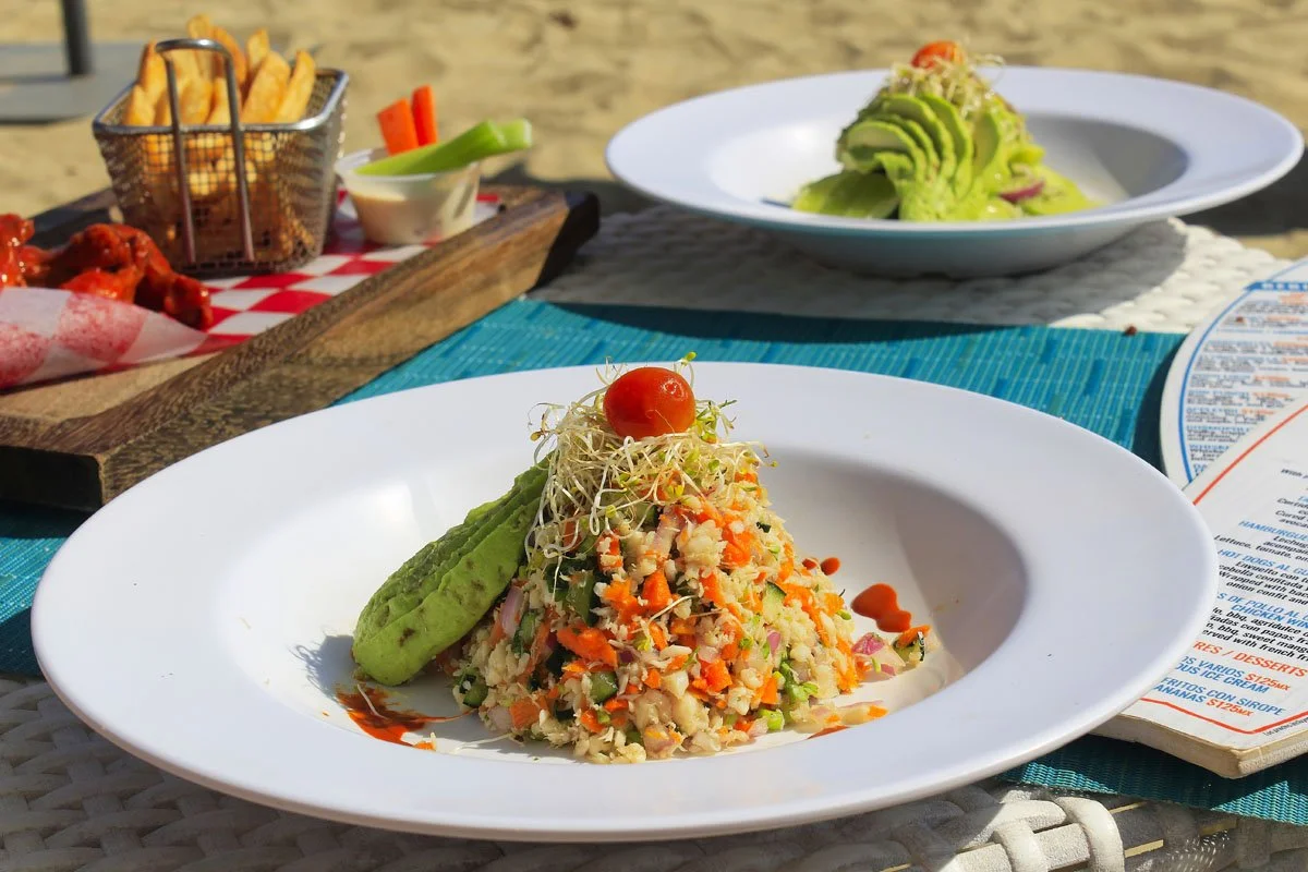 A plate of ceviche with diced vegetables, a cherry tomato garnish, and a side of avocado, on a table at the beach.