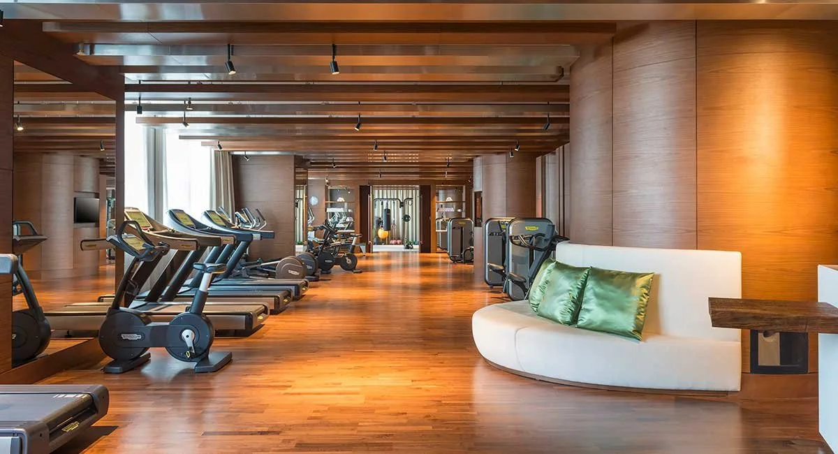 Luxury gym with wood flooring, multiple treadmills and exercise bikes, a white curved sofa with green pillows, and a wooden ceiling with track lighting.